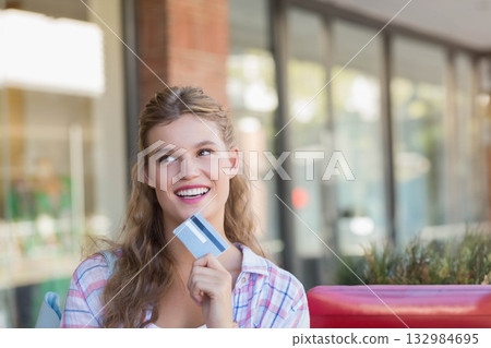 Woman sitting on red bench in front of glass storefront windows holding credit card and smiling 132984695
