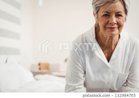 Senior woman sitting on bed edge in lit bedroom wearing pajamas beside white linens, copy space 132984703