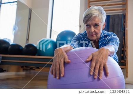 Senior woman leaning forward over purple stability ball inside fitness studio with wall bars 132984831