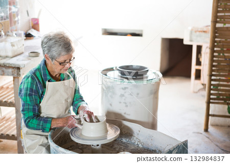 Senior female potter in apron shaping clay vessel on pottery wheel in ceramics studio, copy space 132984837