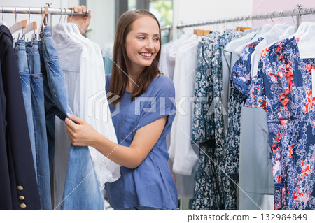 Female customer browsing wooden hanger racks in boutique, holding denim jeans and white blouse 132984849