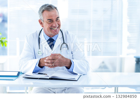 Male physician wearing stethoscope and sitting at clinic desk reading medical textbook with tablet Male physician wearing stethoscope and sitting at clinic desk reading medical textbook with tablet 132984850