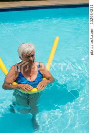 Senior woman wading in backyard pool wearing blue patterned swimsuit holding yellow foam noodle 132984851