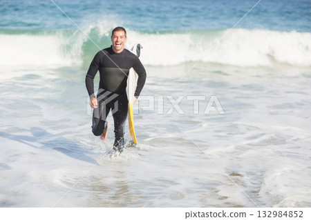 Man emerging from shallow water at shoreline wearing wetsuit and carrying yellow white surfboard Man emerging from shallow water at shoreline wearing wetsuit and carrying yellow white surfboard 132984852