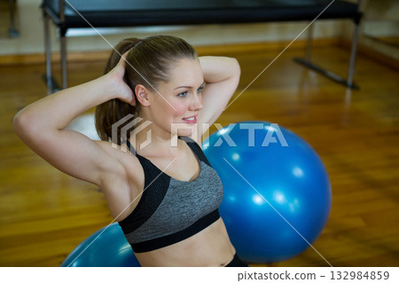 Female athlete balancing core muscles on blue stability ball near workout bench in fitness studio 132984859