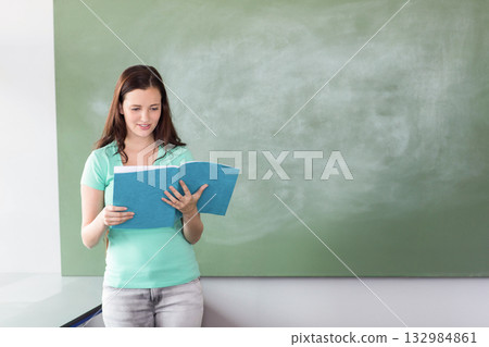 Woman reading from blue binder in classroom before green chalkboard and desk, copy space Woman reading from blue binder in classroom before green chalkboard and desk, copy space 132984861