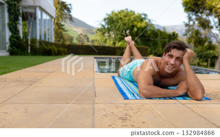 Man lying on striped towel beside pool on tan patio wearing blue swim trunks, copy space 132984866