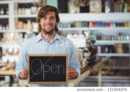 Adult man standing inside cafe holding chalkboard sign while showcasing espresso machine and jars 132984870
