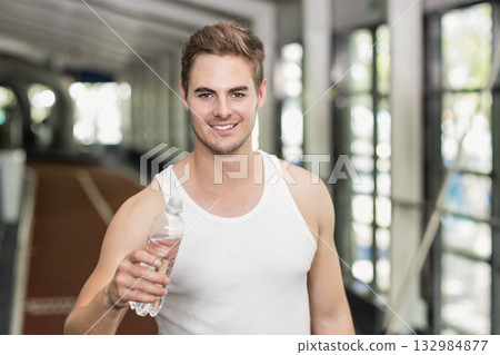 Man standing inside fitness center track corridor wearing white athletic top holding water bottle 132984877