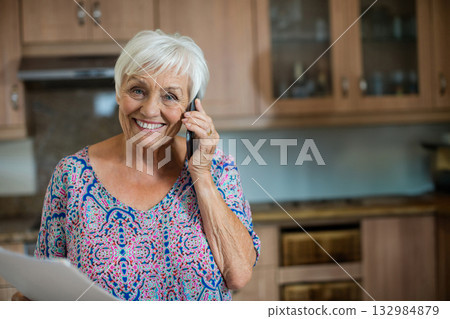 Senior woman standing in home kitchen, holding smartphone and checking papers on countertop Senior woman standing in home kitchen, holding smartphone and checking papers on countertop 132984879