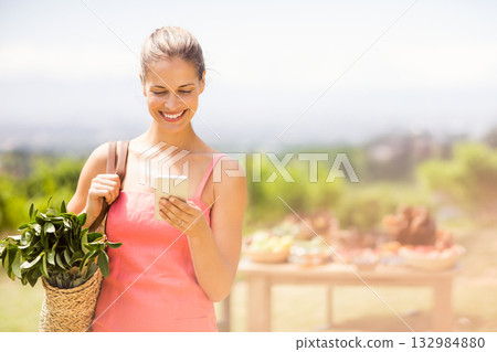 Female shopper holding woven tote bag with potted plant checking smartphone at open-air market Female shopper holding woven tote bag with potted plant checking smartphone at open-air market 132984880