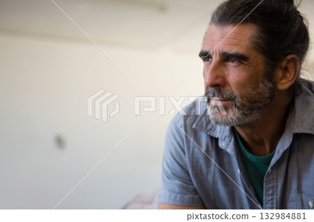 Mature man sitting in home studio gazing toward right side at blank off-white wall, copy space Mature man sitting in home studio gazing toward right side at blank off-white wall, copy space 132984881