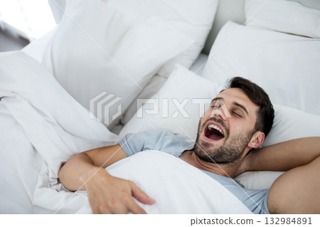 Adult man yawning and stretching upon waking in bedroom on white bed with pillows, copy space 132984891