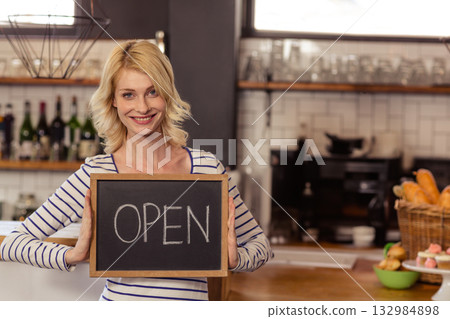 Woman in striped top holding OPEN chalkboard at counter with coffee machine, pastries 132984898
