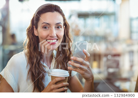 Woman smiling while holding disposable coffee cup and checking smartphone at modern coffee shop 132984900