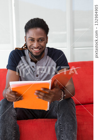 African American man sitting on red sofa reviewing notes in orange binder by white-framed windows 132984905