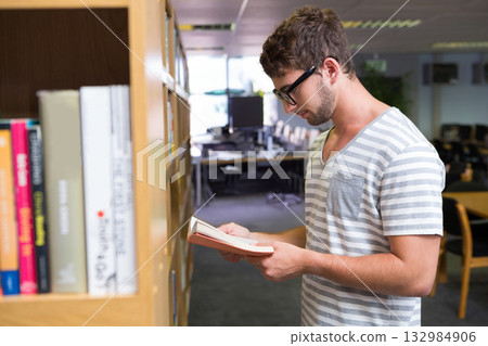 Man standing by bookshelf in library holding open paperback and reading next to computer monitors Man standing by bookshelf in library holding open paperback and reading next to computer monitors 132984906