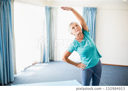 Senior woman in turquoise shirt performing side stretch on blue yoga mat near windows, copy space Senior woman in turquoise shirt performing side stretch on blue yoga mat near windows, copy space 132984913
