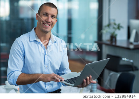 Professional man holding laptop and smiling while standing in open-plan office with desk and plant 132984914