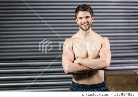 Shirtless athletic man standing with arms crossed in warehouse-style gym by wooden plyometric box 132984922