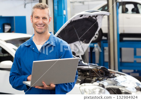 Male mechanic wearing blue coveralls using laptop inspecting lifted car with raised hood in garage 132984924