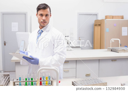 Male scientist recording on clipboard at lab bench near test tubes and microscope Male scientist recording on clipboard at lab bench near test tubes and microscope 132984930