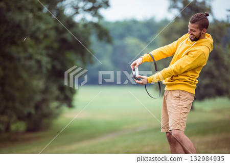 Man wearing yellow hoodie and tan shorts holding film camera smiling in grassy meadow, copy space 132984935