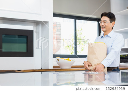 Senior Japanese man holding paper grocery bag with produce beside modern kitchen island, copy space 132984939