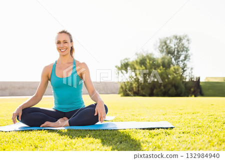 Woman in teal tank top and navy leggings sitting cross-legged on blue yoga mat in garden 132984940