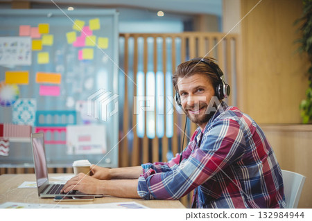 Mid adult man with headphones using laptop with coffee at office desk beside whiteboard, copy space 132984944