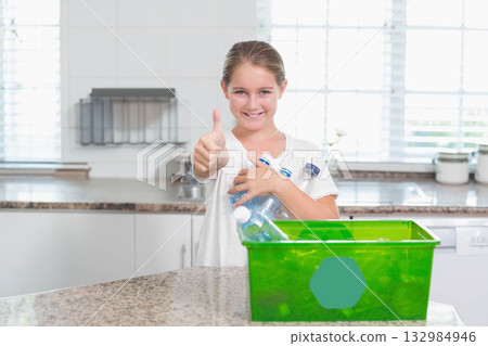 Girl standing at countertop holding empty bottles near green recycle bin giving thumbs up at home 132984946