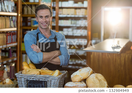 Male shopkeeper wearing apron standing behind basket of baguettes and loaves on counter in bakery 132984949