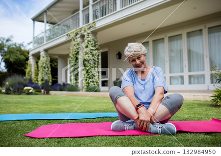 Senior woman sitting crosslegged on pink and blue mats on home lawn wearing sneakers and smartwatch Senior woman sitting crosslegged on pink and blue mats on home lawn wearing sneakers and smartwatch 132984950