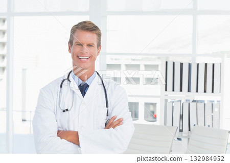 Male doctor standing in exam room wearing lab coat with stethoscope beside binder-filled shelves Male doctor standing in exam room wearing lab coat with stethoscope beside binder-filled shelves 132984952