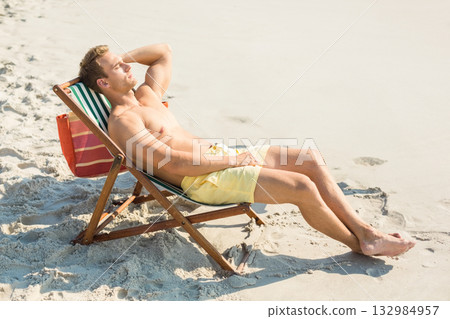 Shirtless man reclining in beach chair on beach wearing yellow swim trunks beside striped tote bag Shirtless man reclining in beach chair on beach wearing yellow swim trunks beside striped tote bag 132984957