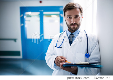 Male doctor leaning on wall in hospital corridor holding blue clipboard and stethoscope, copy space 132984968
