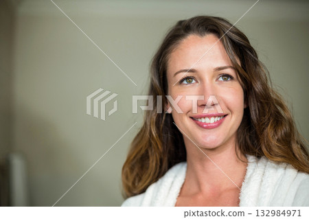 Woman standing against beige bathroom wall wearing white plush bathrobe and smiling, copy space 132984971