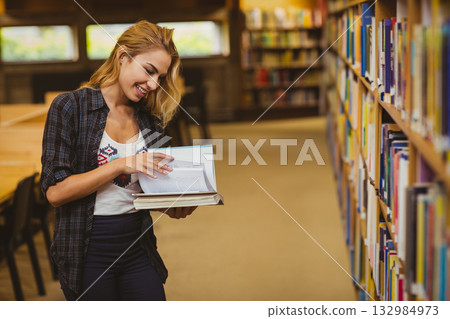 Woman in her twenties holding two books flipping one open beside bookshelf at library, copy space 132984973