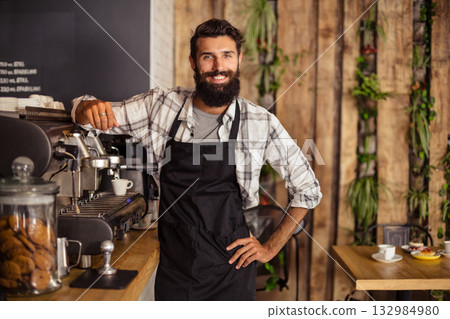 Bearded man wearing black apron leaning on espresso maker at coffee shop counter with cookie jar Bearded man wearing black apron leaning on espresso maker at coffee shop counter with cookie jar 132984980
