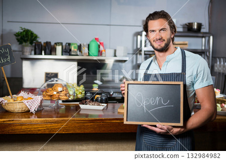 Man in apron holding chalkboard sign showing Open behind bakery counter with pastries, copy space Man in apron holding chalkboard sign showing Open behind bakery counter with pastries, copy space 132984982