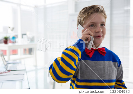 Male child holding smartphone to ear wearing striped sweater, red bow tie in office with laptop 132984983