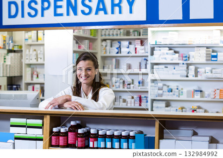 Female pharmacist leaning forward behind wooden dispensary counter, displaying prescription bottles 132984992