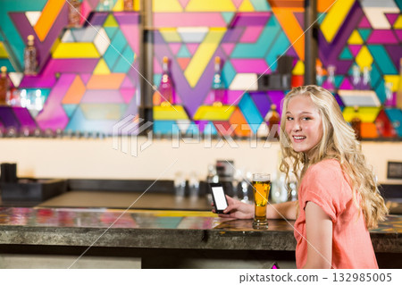 Teenage girl holding smartphone and sipping beer at pub counter with liquor shelves, copy space 132985005