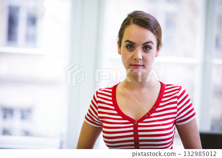 Woman standing facing camera by floor-to-ceiling windows in bright studio wearing striped shirt Woman standing facing camera by floor-to-ceiling windows in bright studio wearing striped shirt 132985012
