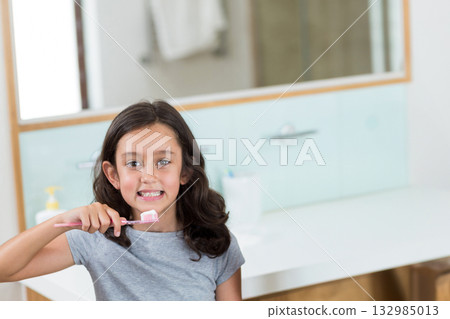 School-age girl brushing teeth at home bathroom vanity using pink toothbrush near soap dispenser 132985013