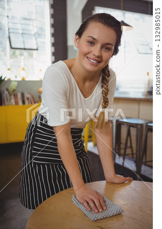Female cafe server wearing striped apron leaning over table wiping with cloth in cafe by windows 132985015
