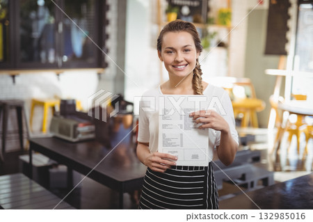 Smiling female server in striped apron holding printed menu at cafe with potted plants 132985016