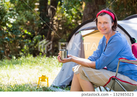Senior woman sitting on chair holding mug beside tent with lantern at forest clearing, copy space Senior woman sitting on chair holding mug beside tent with lantern at forest clearing, copy space 132985023