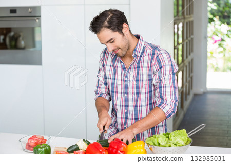 Man slicing zucchini with knife on counter in home kitchen with bell peppers, tomatoes, copy space 132985031
