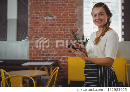 Female server wearing striped apron holding tablet in cafe near brick wall and tables, copy space 132985035